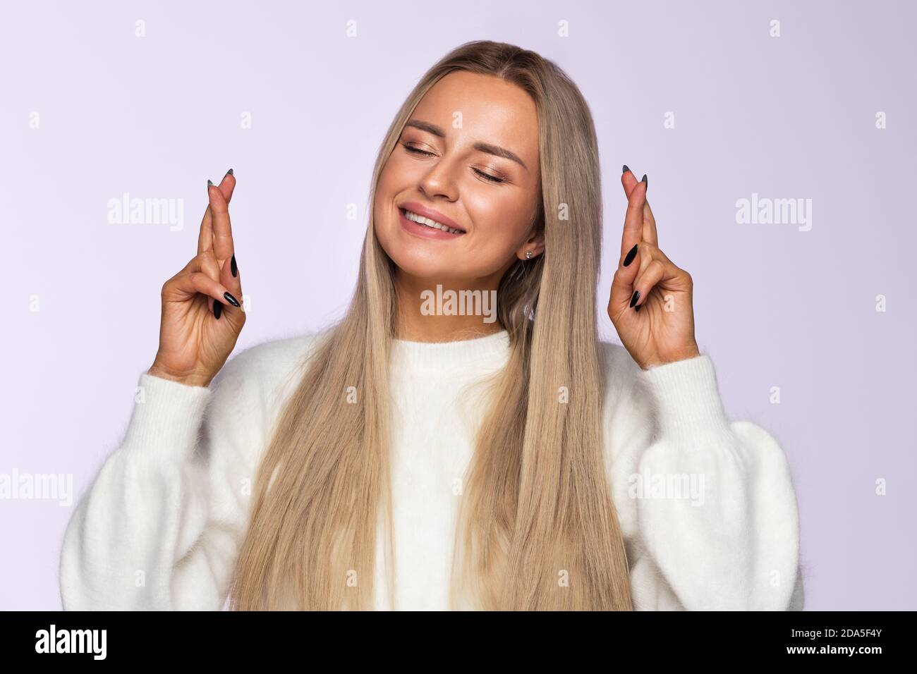 Closeup portrait of positive woman crossing her fingers for good luck and closed eyes, wishful thinking, charming smile, isolated on soft violet backg Stock Photo