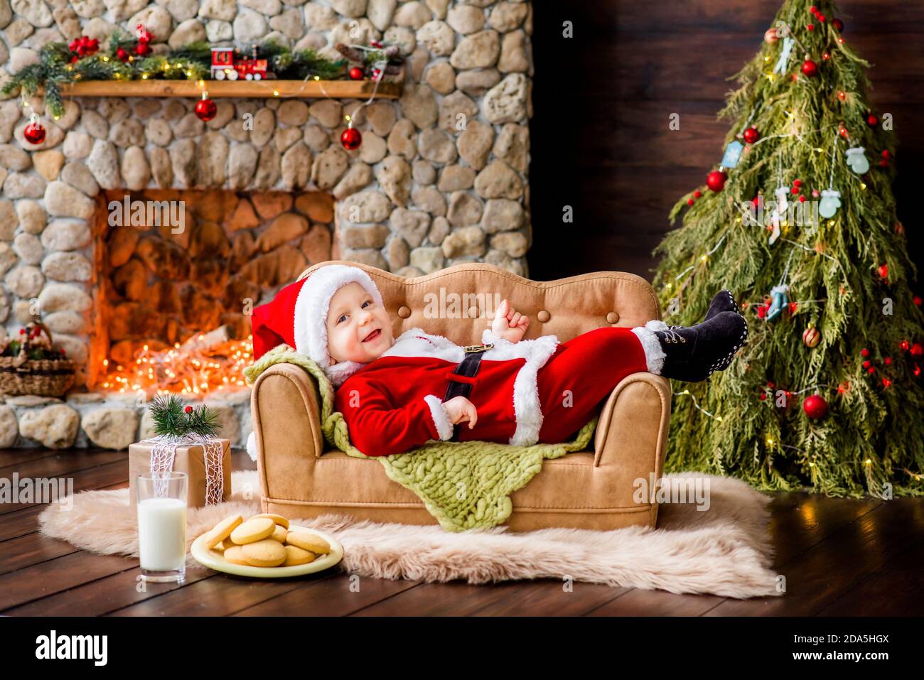 Close-up portrait of a Little boy dressed as red Santa Claus, playing by stone fireplace with bright garlands, putting a gift box under Christmas tree Stock Photo
