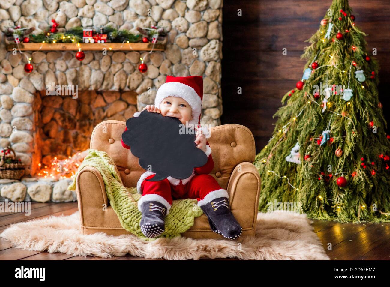 A small child in a red Santa Claus costume, sitting on the couch in the  residence, holds a black cloud plate for text in his hands. New Year card  Stock Photo -