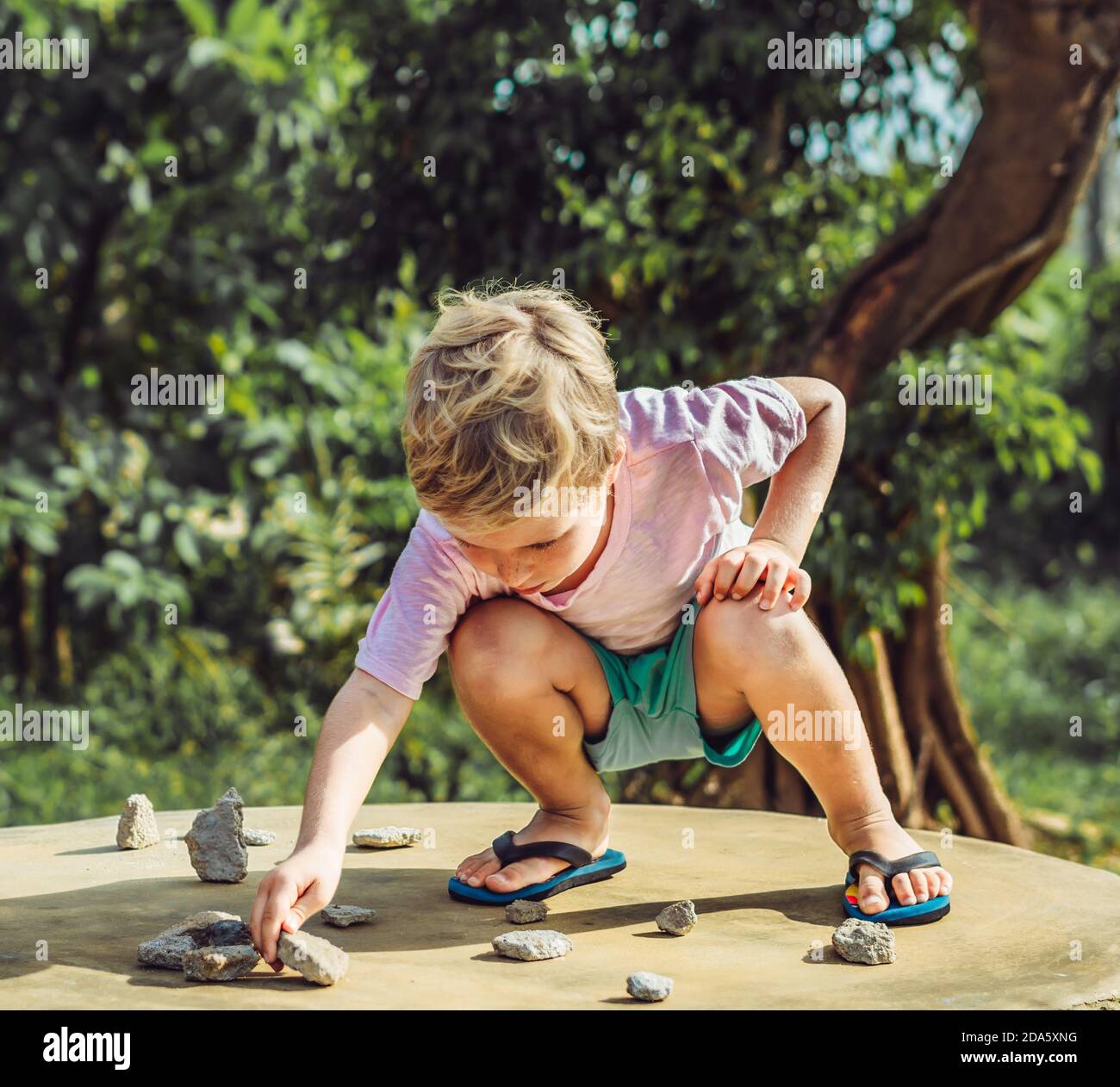 Cute Child kid blond freckled face boy play squatting outside nature sun day, driving traffic regulation rules use stone imitation cars. Expressive Stock Photo
