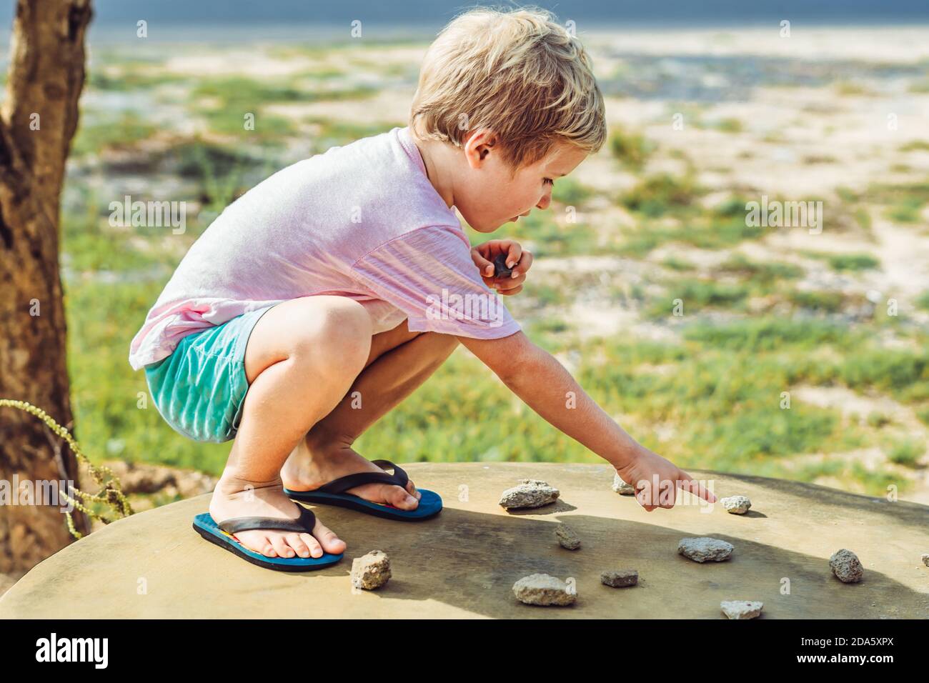 Cute Child freckled face boy play squatting outside nature sun day, driving traffic regulation rules use stone imitation cars toy. Expressive Serious Stock Photo