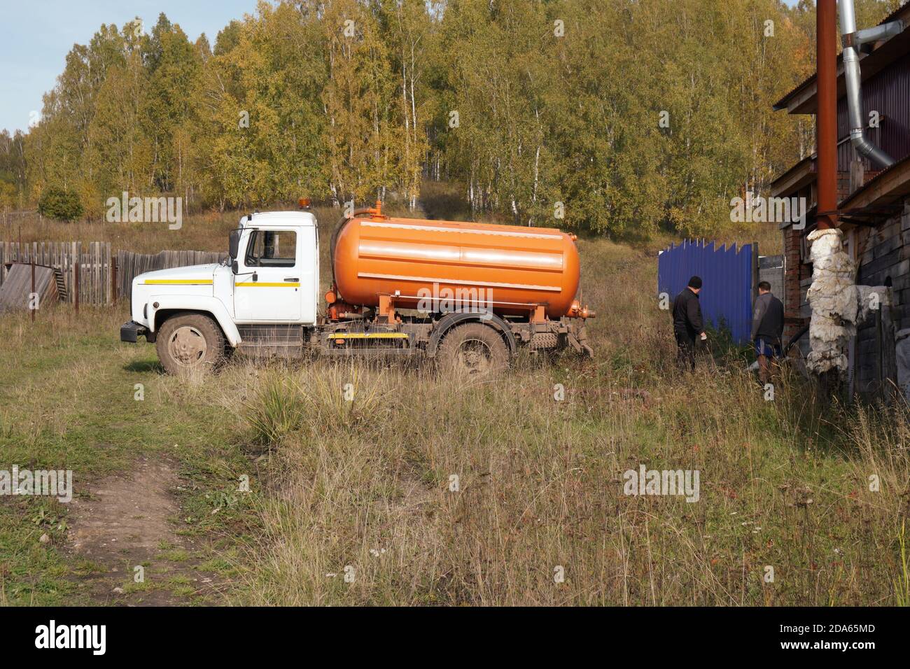 A cesspool machine and two men are standing near a village building amid an autumn birch grove. Stock Photo