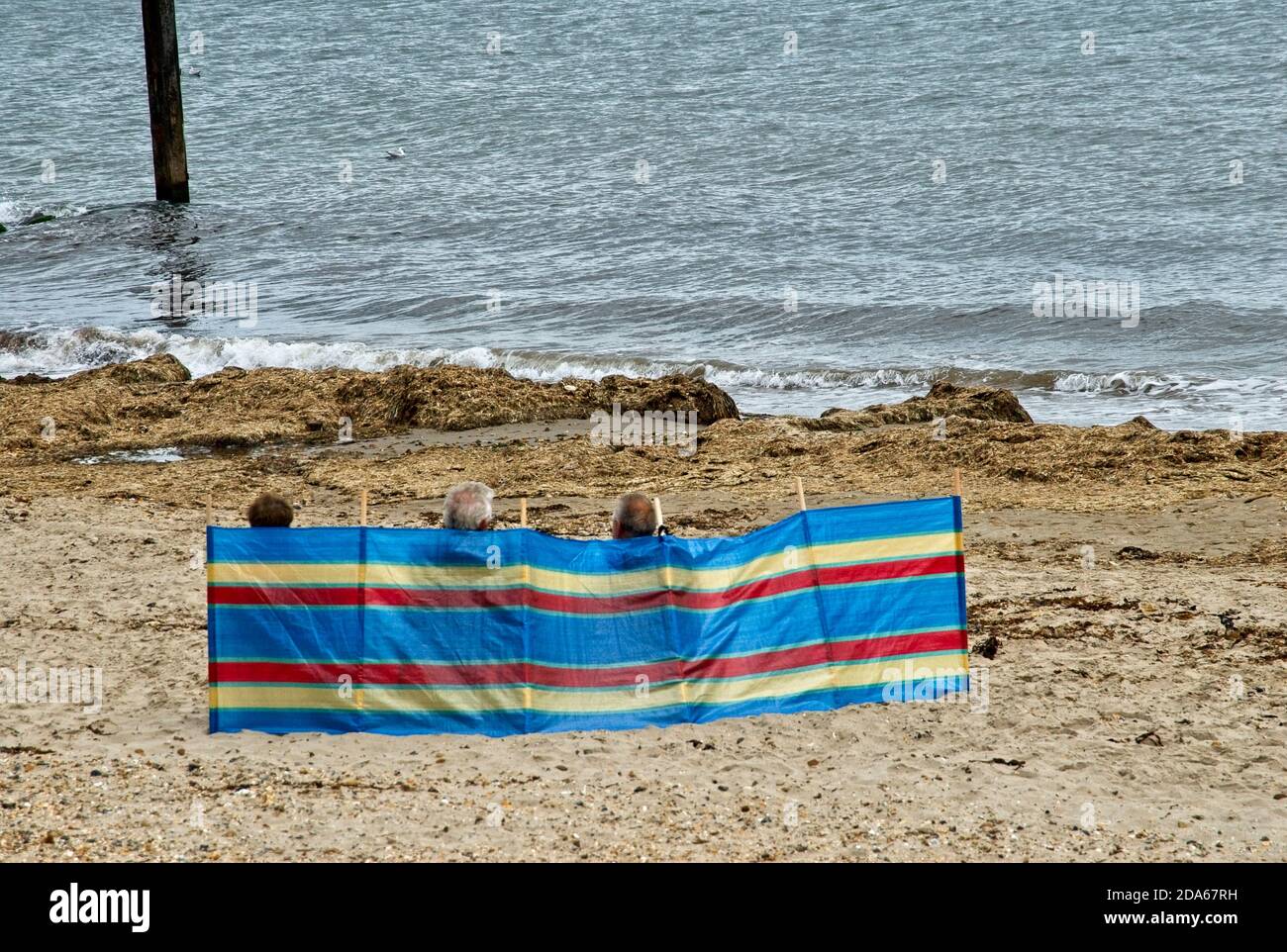 Holiday makers braving cold weather to sit on beach behind windbreak Stock Photo