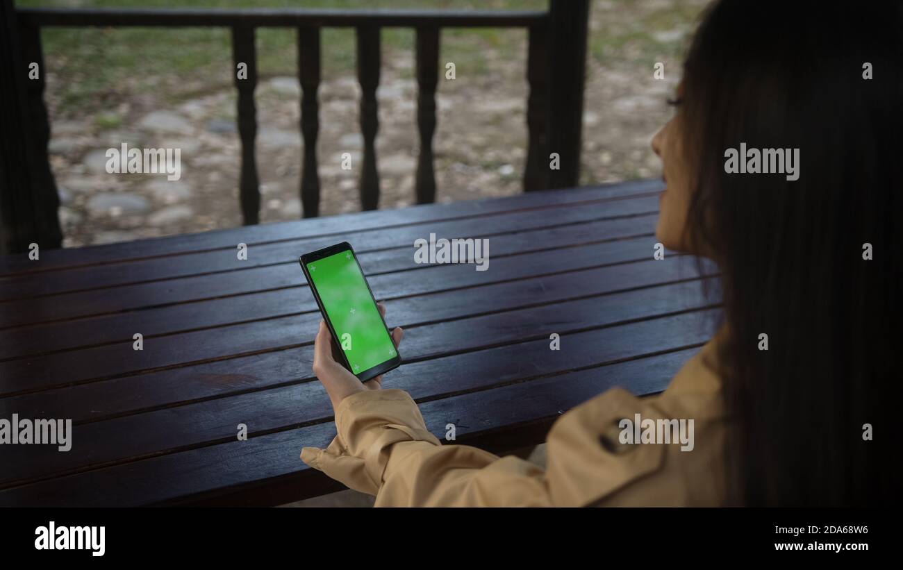 Young woman at nature sitting on a bench using with Green Mock-up Screen Smartphone in Horizontal Landscape Mode. Girl Using Mobile Phone, Browsing In Stock Photo