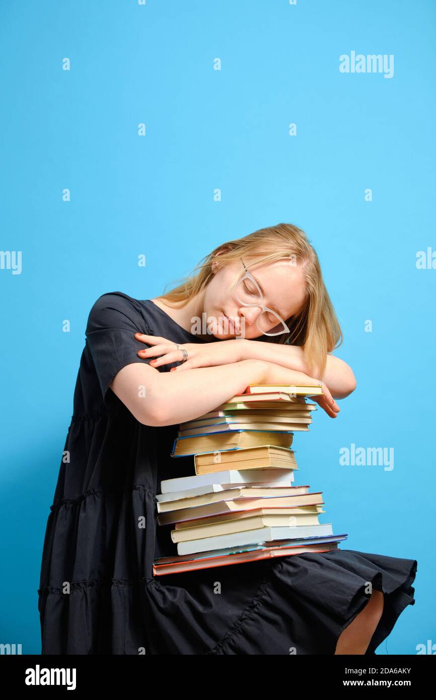 Tired Caucasian girl student laid her head and sleeps on a stack of books on a blue background. Vertical shot and copy space. Stock Photo