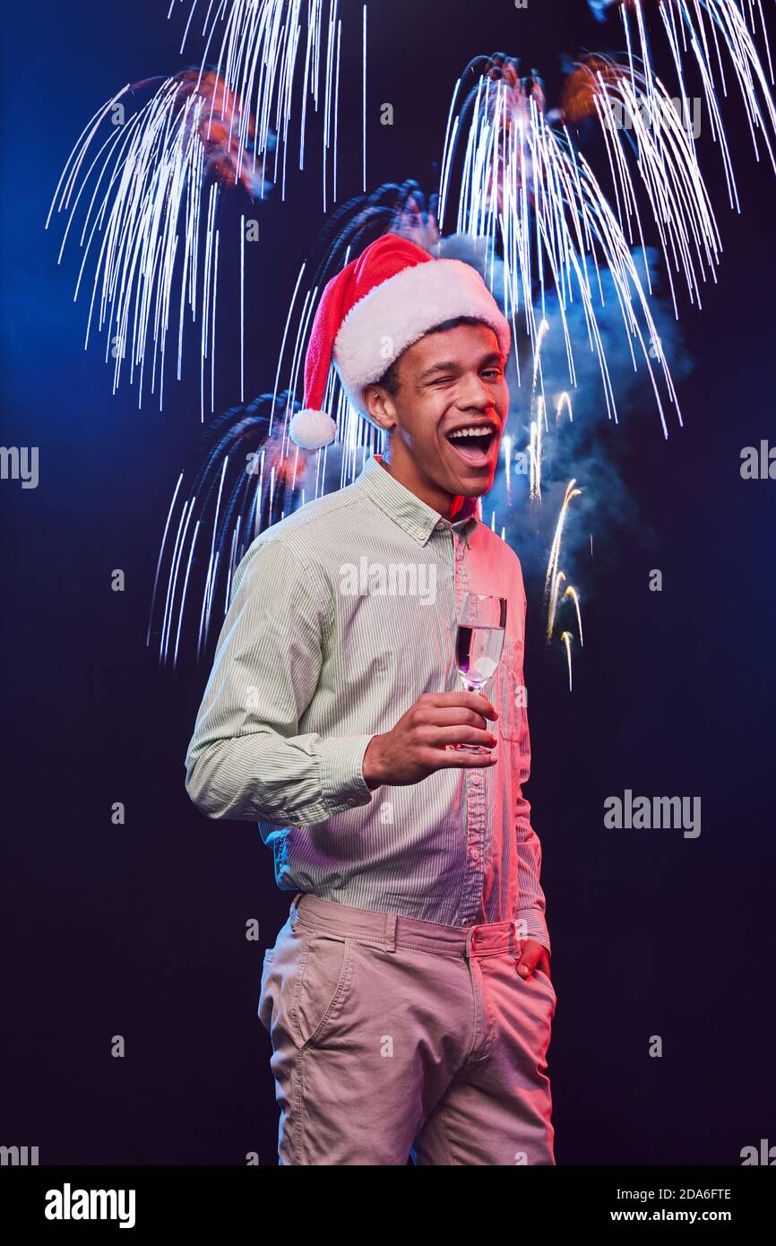 Marry Christmas. Vertical shot of a young excited mixed race man wearing Santa Claus cap with glass of champagne standing against fireworks background. People and holidays Stock Photo