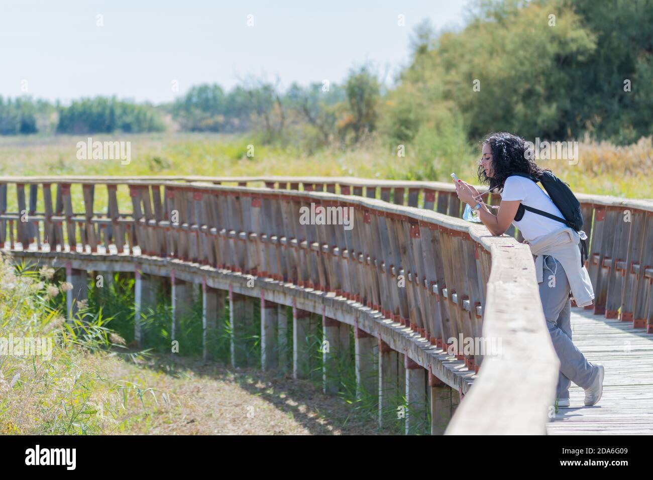 young hiker woman with curly black hair looking at her cellphone while taking a walk on a wooden bridge on a sunny day Stock Photo