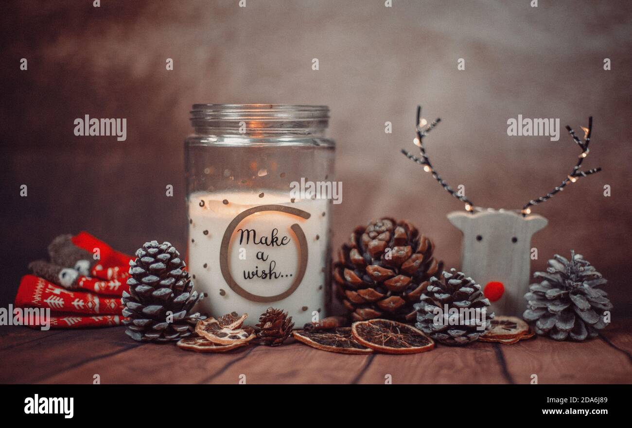 Christmas composition with 'make a wish' candle in a glass jar with rain deer, Christmas lights and pine cones on a rustic brown background. Christmas Stock Photo