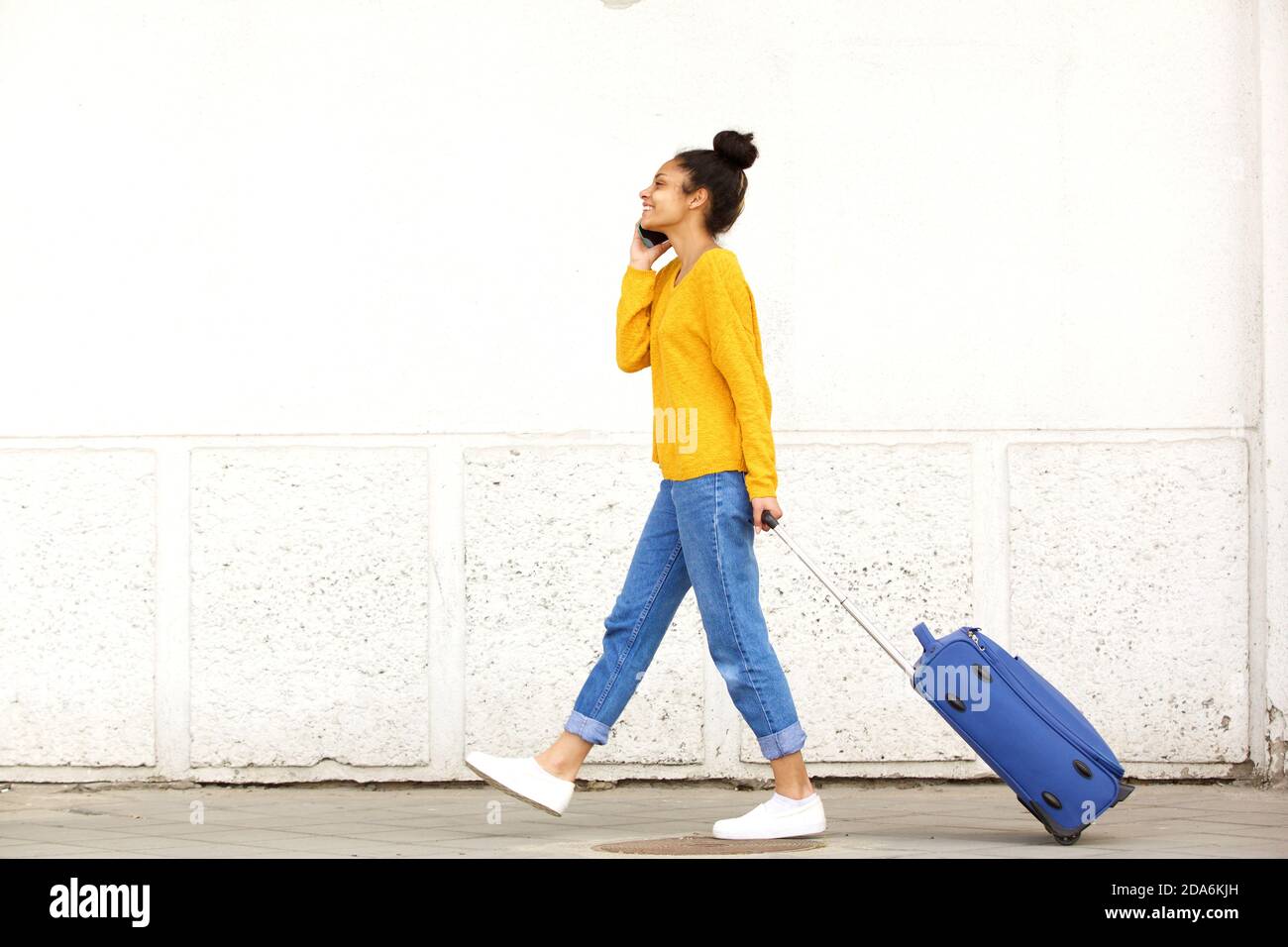 Full body side view portrait of young woman traveling with suitcase and talking on mobile phone
