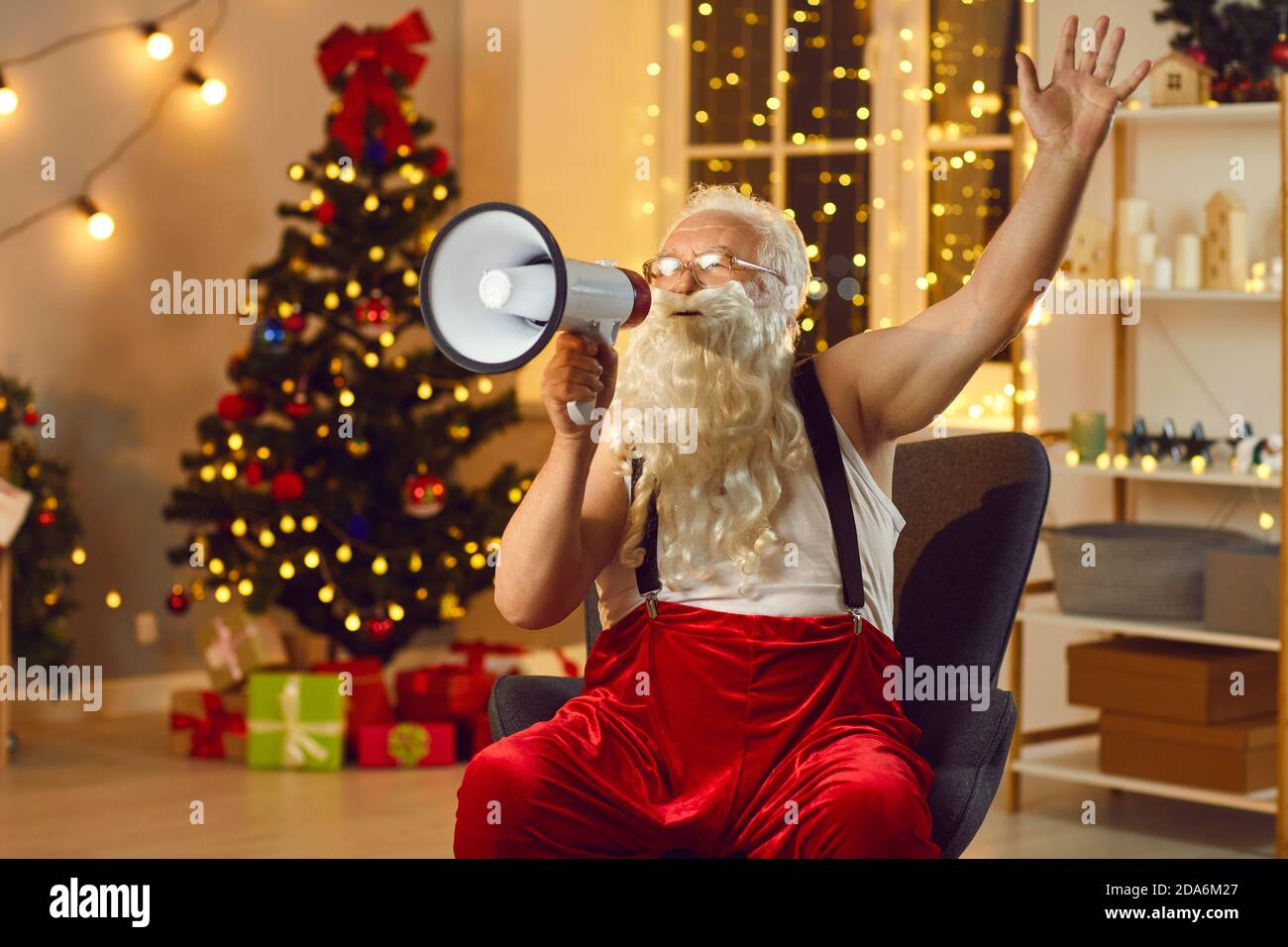 Senior Santa Claus speaks into a loudspeaker while sitting in a room with Christmas decorations. Stock Photo