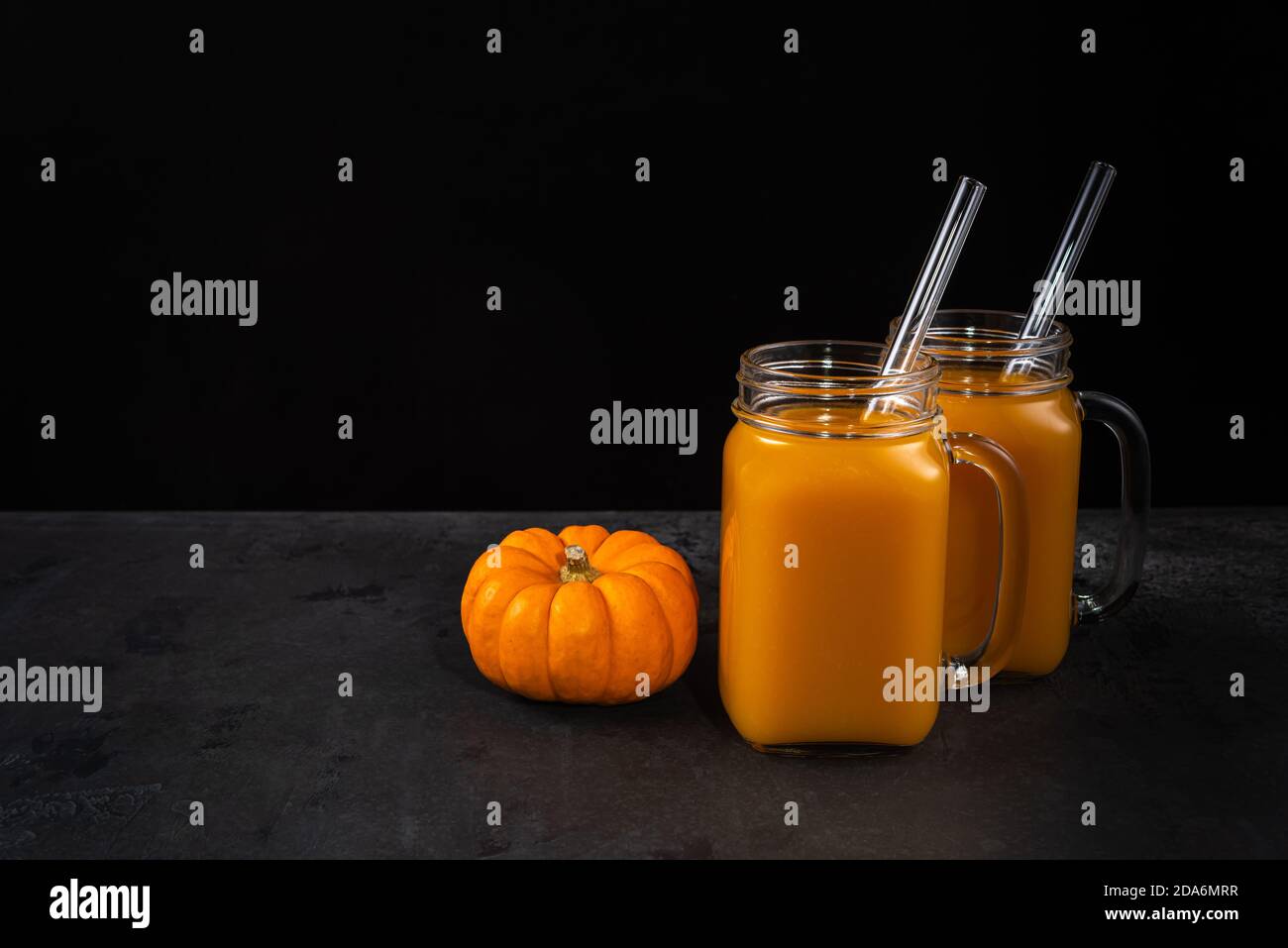 Whole pumpkin, square glass jars with non-alcoholic mocktail and glass straws on black textured surface. Horizontal background for Halloween with copy Stock Photo