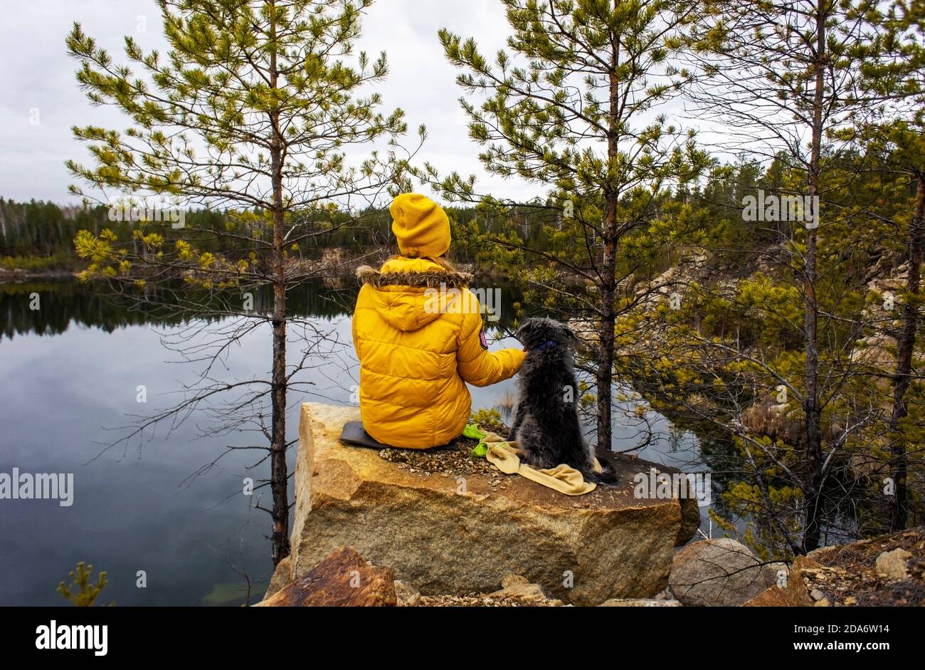 Young woman in yellow clothes from the back with a fluffy gray dog sitting on a rocky lake near the pine trees. Autumn beautiful landscape, natural ba Stock Photo