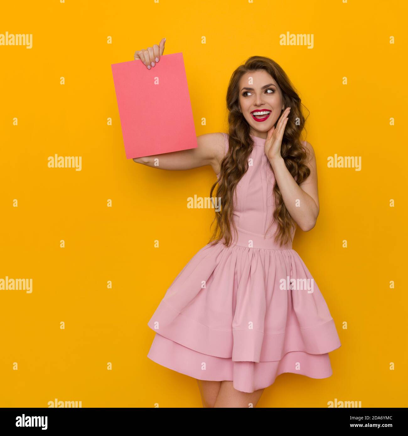Happy beautiful young woman in pink cocktail dress is holding pink empty sheet of paper and talking. Front view. Three quarter length studio shot on y Stock Photo