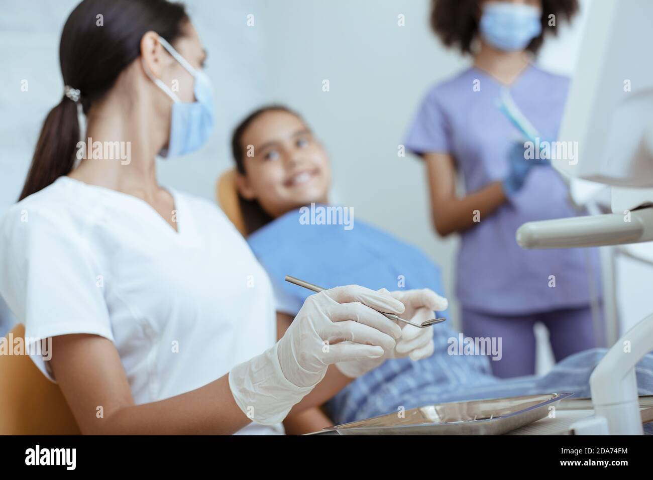 Young lady doctor in uniform, rubber gloves and mask with tool treats tooth of smiling little child Stock Photo