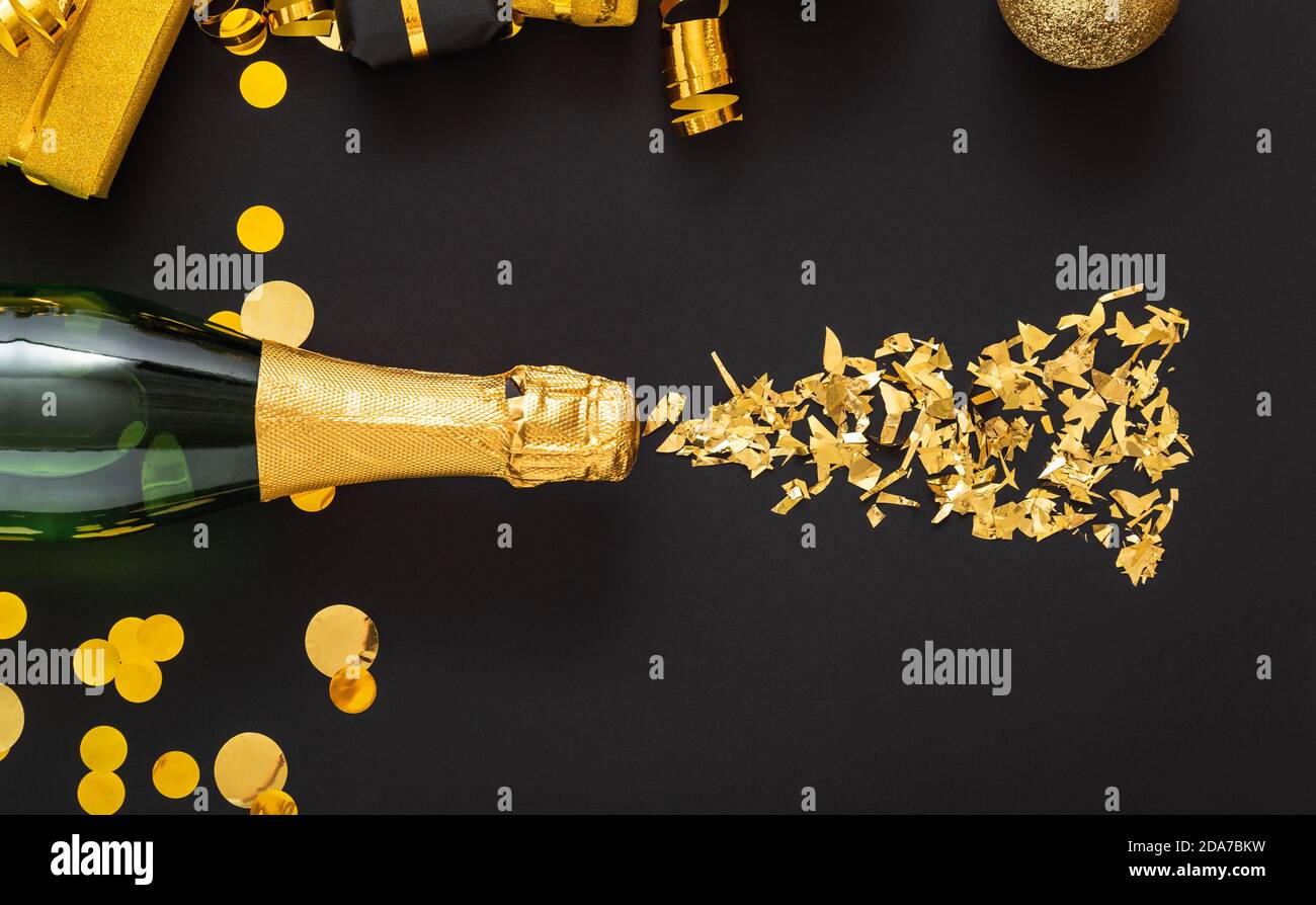 Golden bottle of champagne spills out gold sparkles in frame of gold festive Christmas decor confetti balls gifts on black background. Flat lay New Ye Stock Photo