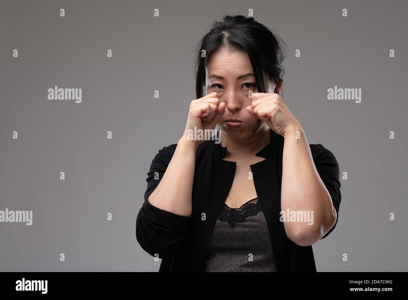 Upset morose Asian woman standing crying raising her hands to her eyes in sorrow over a grey studio background with copyspace Stock Photo