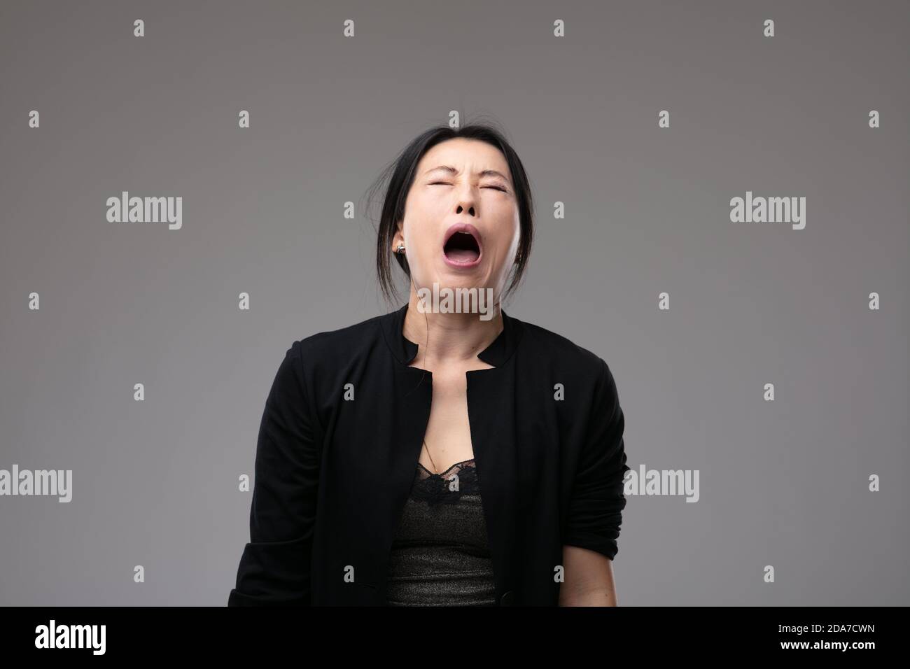 Woeful Asian woman wailing in anguish with her mouth open and a sorrowful expression over a grey studio background with copyspace Stock Photo