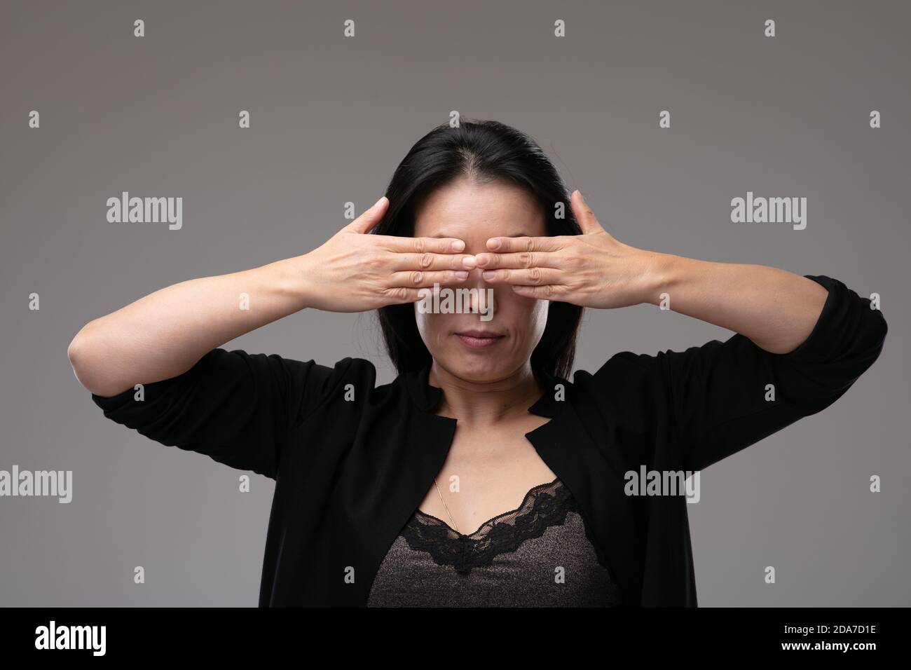 Asian woman in a smart black outfit covering her yes with her hands in a concept of the metaphor see no evil, hear no evil speak no evil, over a grey Stock Photo