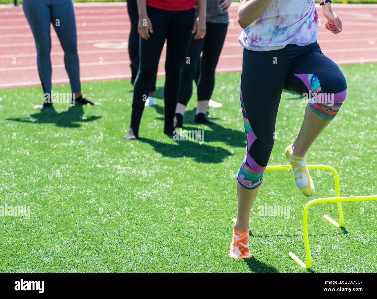 Track runners stepping over yellow mini hurdles with no shoes on wearing socks on a green turf field during high school track and field practice. Stock Photo