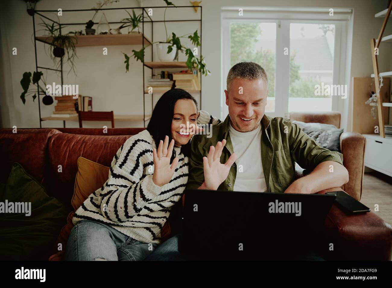 Cheerful couple sitting on sofa and waving hands to laptop while chatting using video call at home Stock Photo