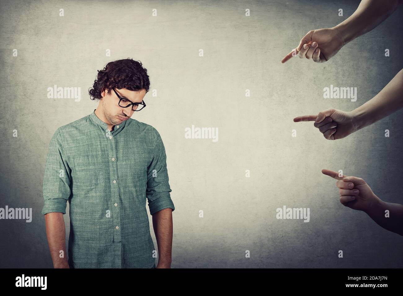 Stressed man, curly hair wears eyeglasses, looks down exhausted and depressed, feels ashamed being under pressure, as multiple hands pointing forefing Stock Photo