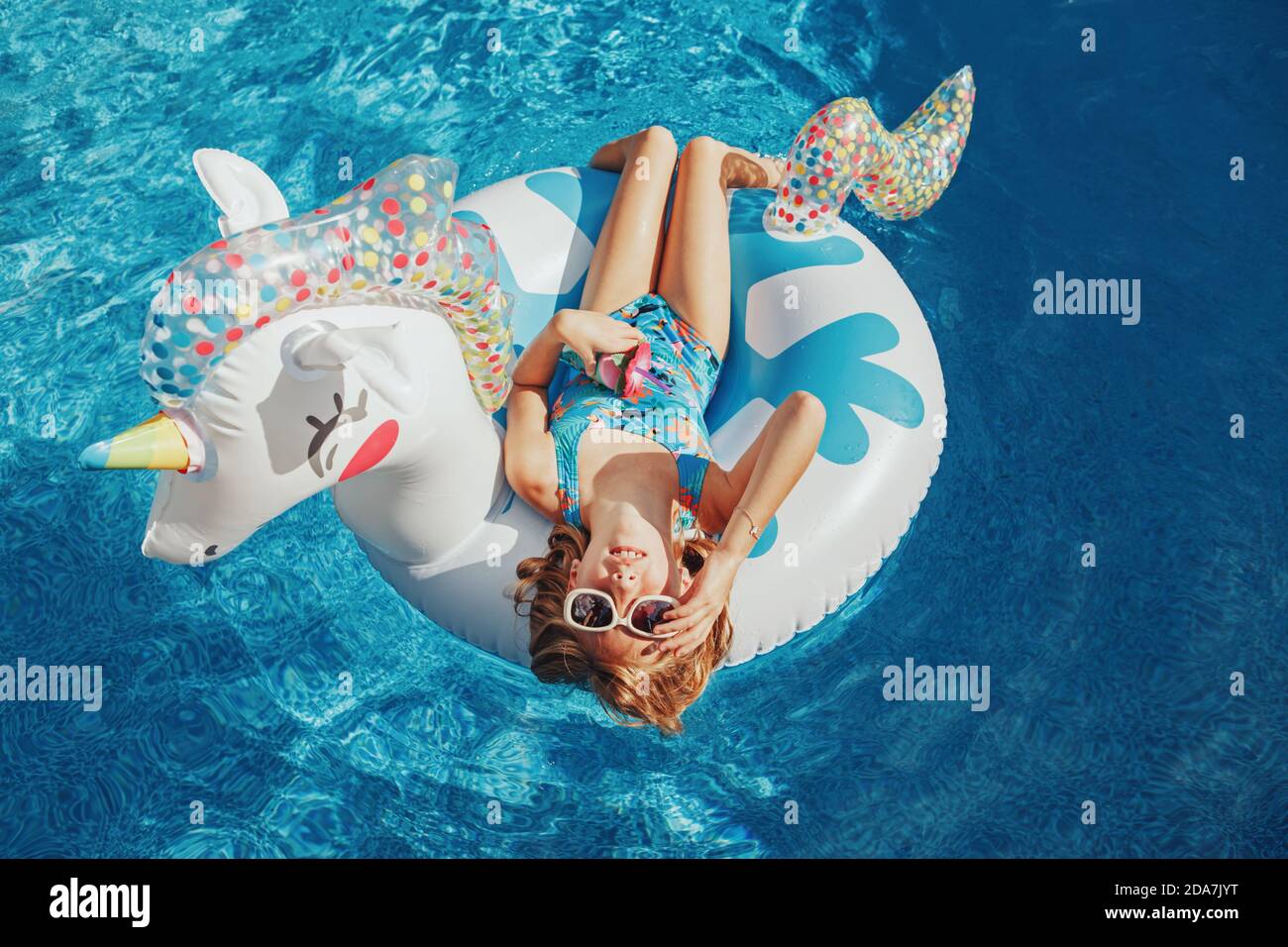 Cute adorable girl in sunglasses with drink lying on inflatable ring unicorn. Kid child enjoying having fun in swimming pool. Summer outdoor water Stock Photo