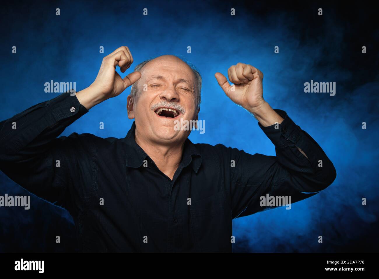 Old macho man. An elderly man holds his hands behind his head on a dark ...