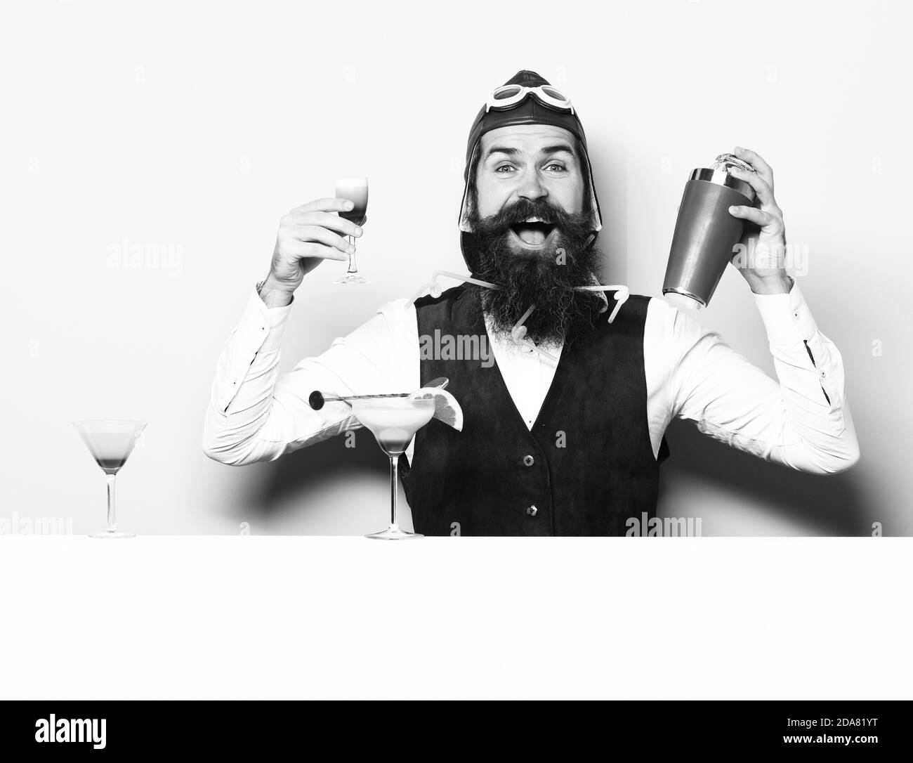 handsome bearded aviator or pilot with long beard and mustache on smiling face with shaker alcoholic cocktail in vintage suede leather waistcoat with hand and glasses on purple green studio background Stock Photo