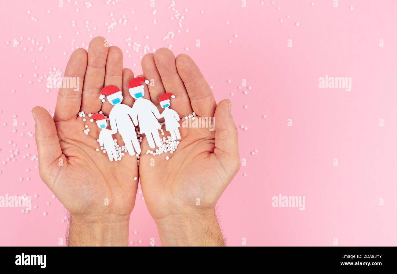 Hands holding a family cut out of paper with a mask and Christmas hat on a pink background. Stock Photo