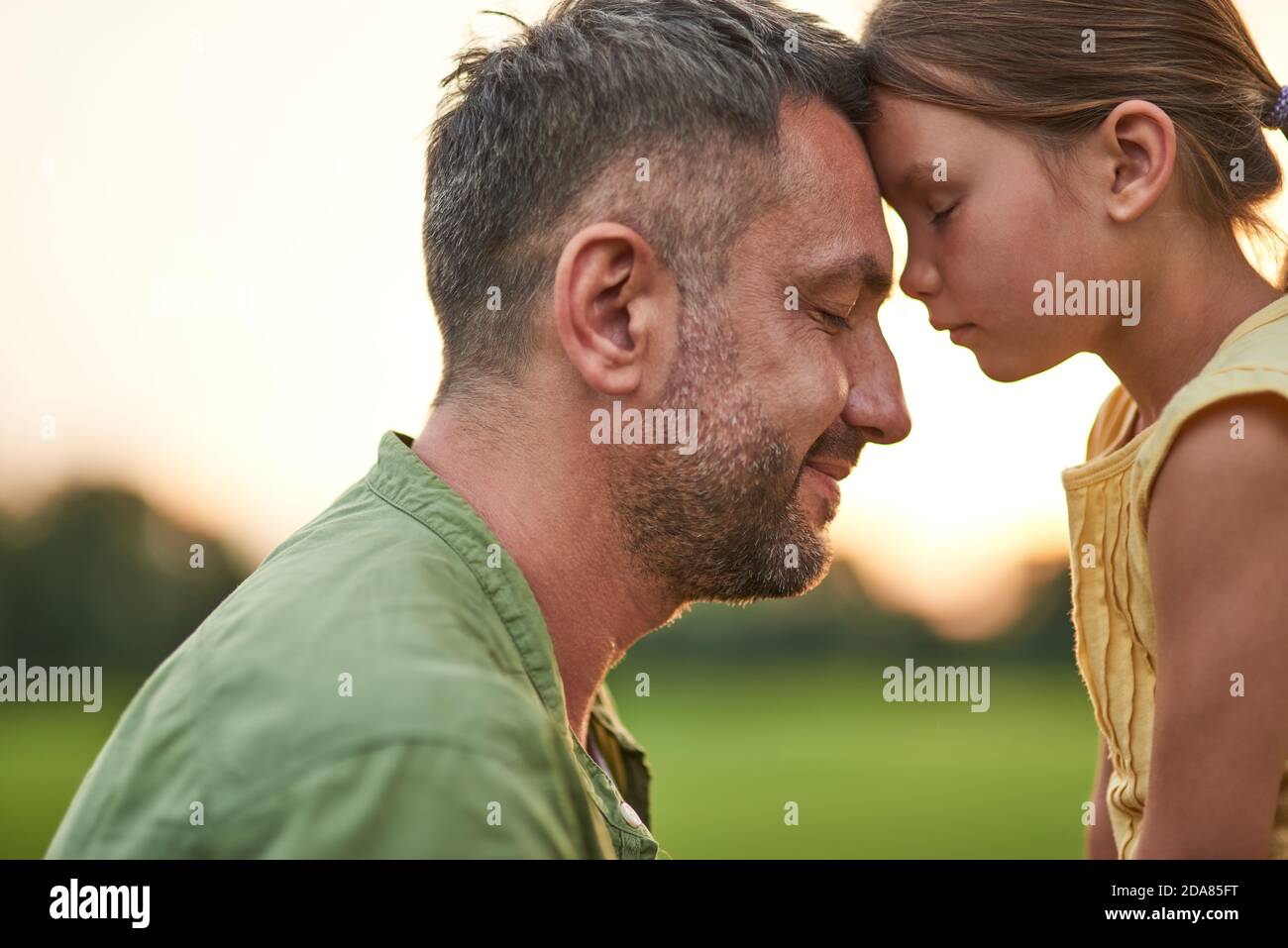 Close up shot of young father and adorable little daughter touching foreheads, standing with eyes closed while spending time outdoors in the park on a Stock Photo