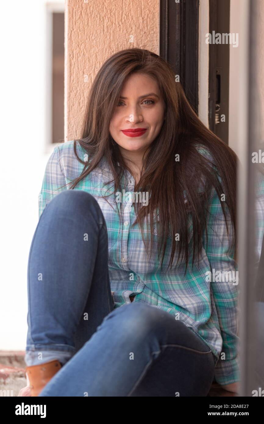 attractive young latin woman with long hair and makeup, happy and sitting on the edge of the window, wearing a plaid t-shirt and jeans Stock Photo