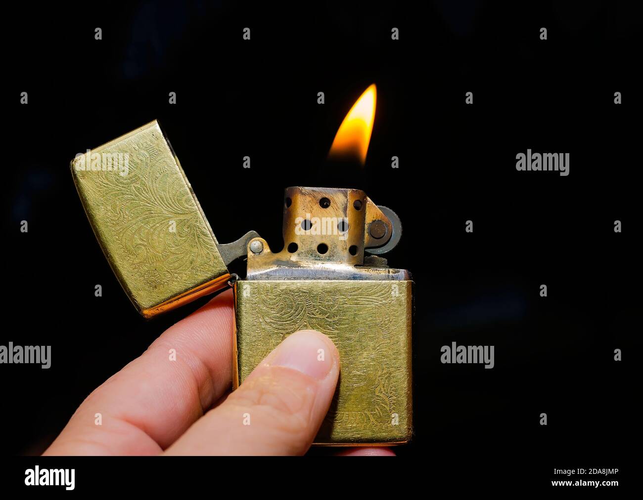 A hand holds a Venetian gold Zippo lighter open and lit. Zippo lighters are reusable metal lighters manufactured by American Zippo Manufacturing. Stock Photo