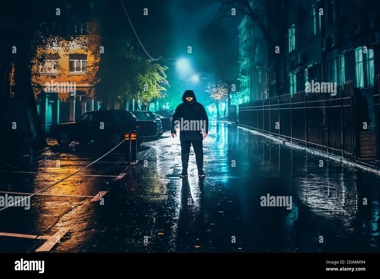 Lonely man in hood standing under rain on empty wet street in autumn night, evening city lights reflection in puddles on asphalt. Stock Photo