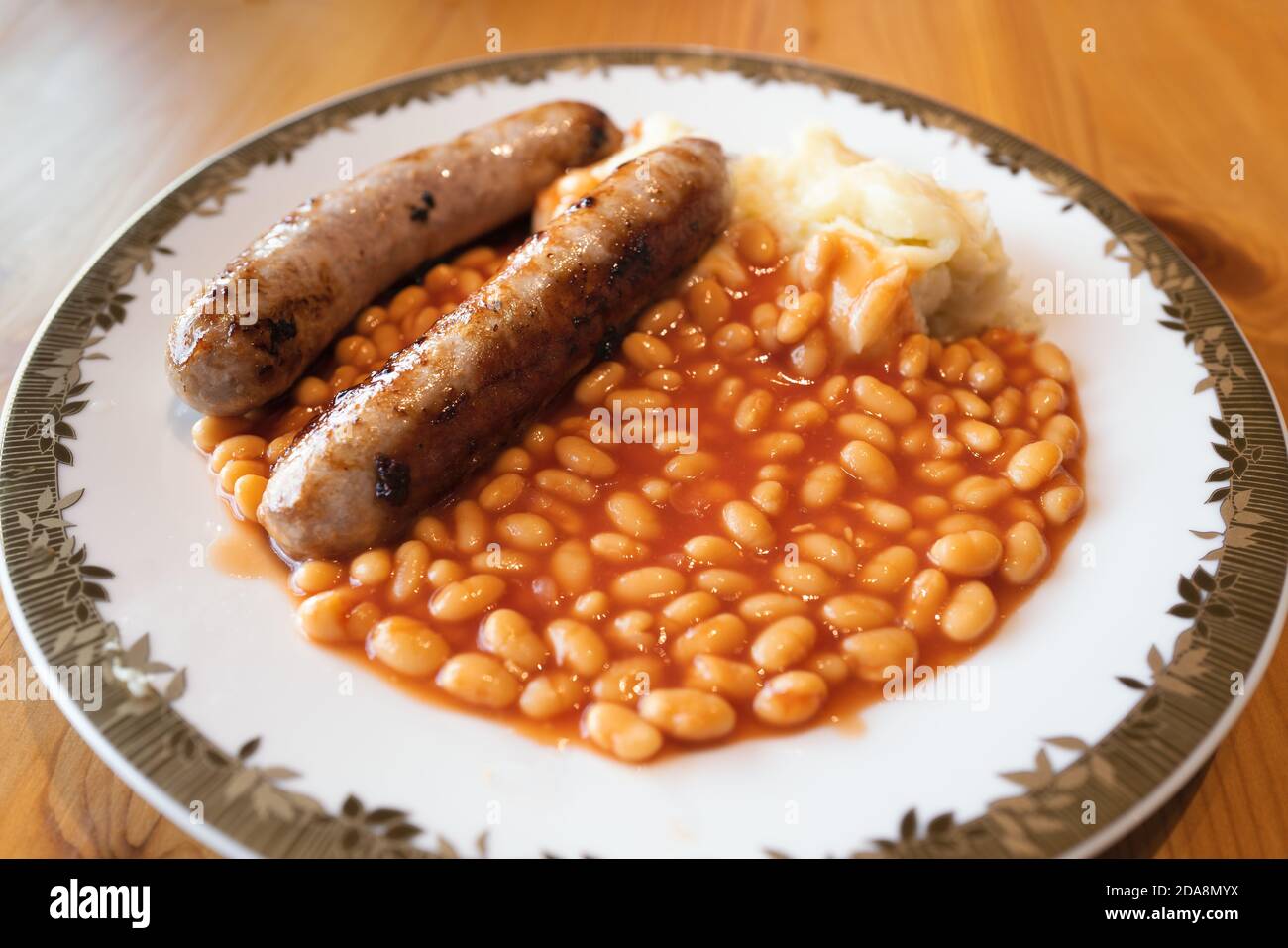 Two large fried sausages with baked beans and mashed potato on a white plate with a gold colour trim on a wooden table lit by window natural light. Stock Photo