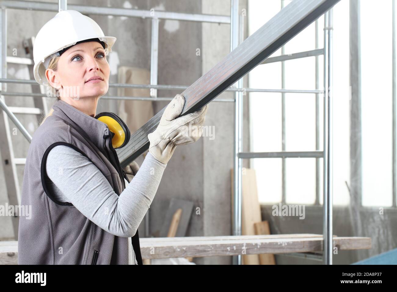 woman construction worker builder portrait wearing white helmet and hearing protection headphones, holding a metal stud for drywall on interior site b Stock Photo