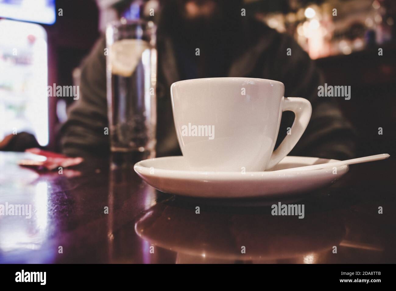 A cup full of coffee or tea on the wooden table in a bar. A man and a glass of mineral water in the background. Stock Photo