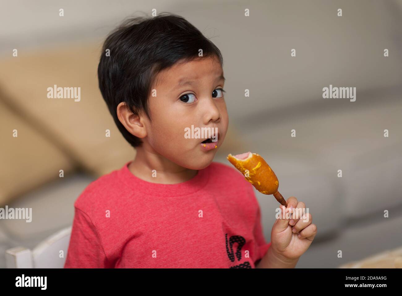 A young boy looks with awe as he takes a bite out of a homemade corn dog, a healthy snack. Stock Photo
