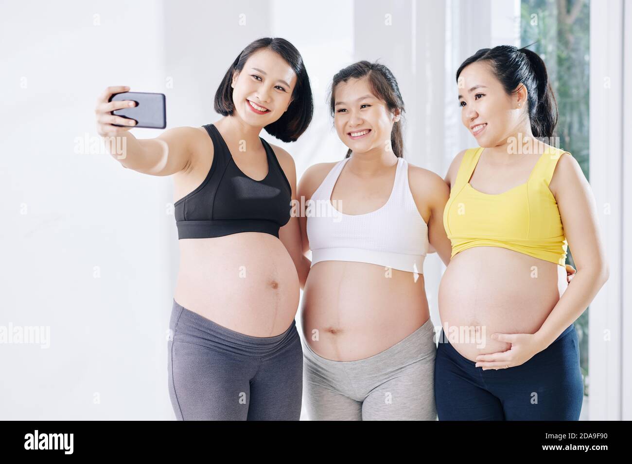 Pretty smiling pregnant Asian woman taking selfie with other pregnant women after yoga class Stock Photo