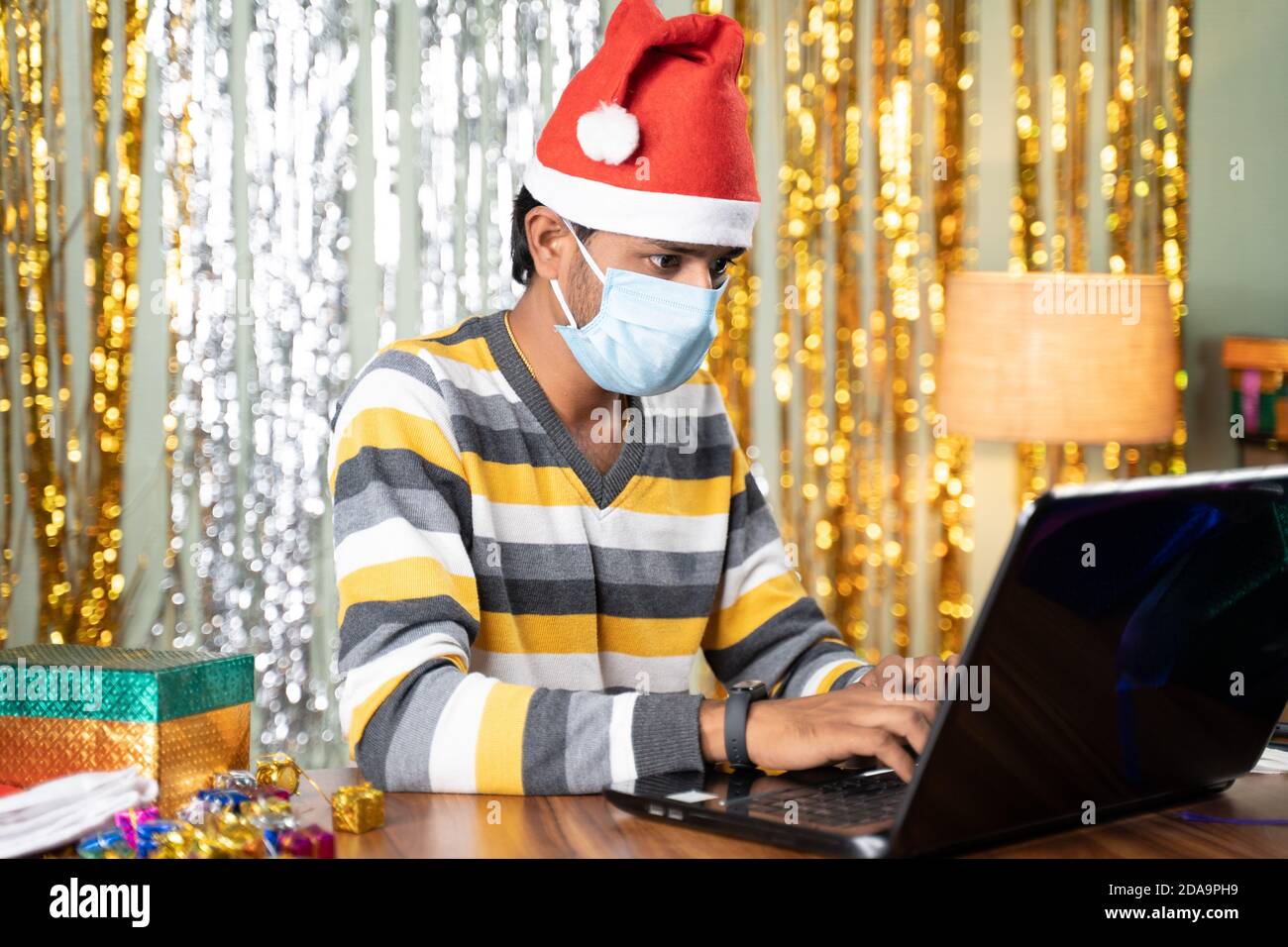 Young man in medical mask busy working on laptop during Christmas or new year celebration eve with decorated background and gifts in front - concept Stock Photo