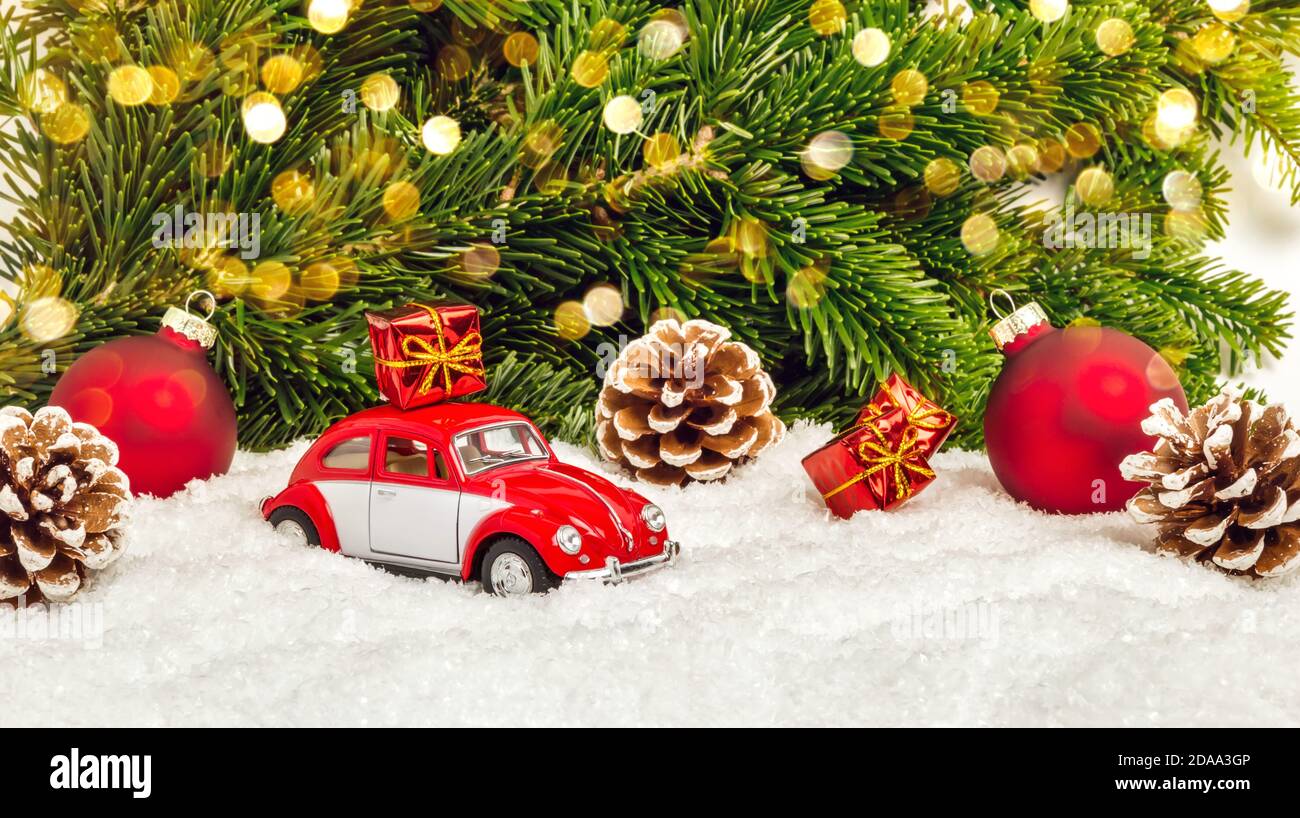 Fulda, Germany - October 24, 2020: Red car in snow with pine cones, Christmas baubles, gifts, fir tree and bokeh lights. Winter holiday trip or delive Stock Photo
