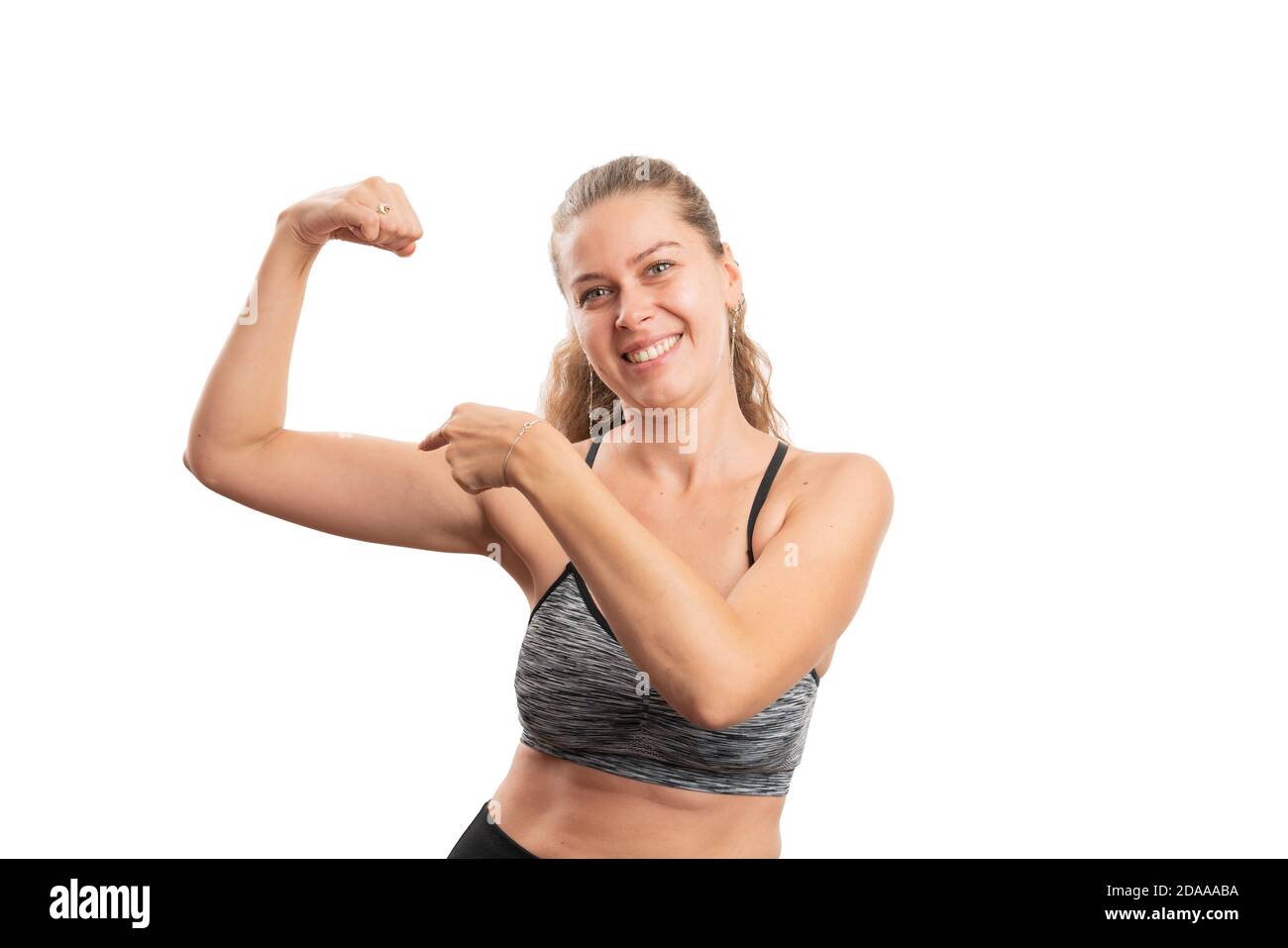 Cheerful woman model pointing index finger at biceps muscle as healthy workout concept wearing sports gym attire isolated on white studio background w Stock Photo