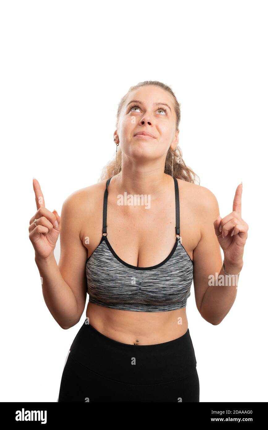 Friendly fit adult woman wearing sports training attire pointing index fingers at copyspace above head as fitness lifestyle concept isolated on white Stock Photo