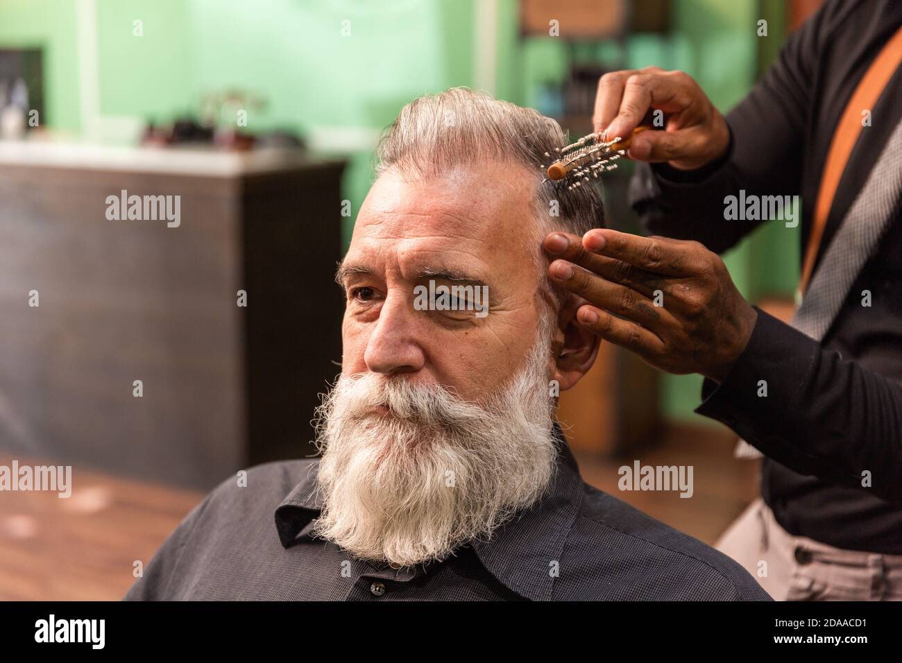young african american barber and dreadlocks, with coronavirus pandemic prevention mask, combs with a comb and his hands a mature white caucasian man Stock Photo