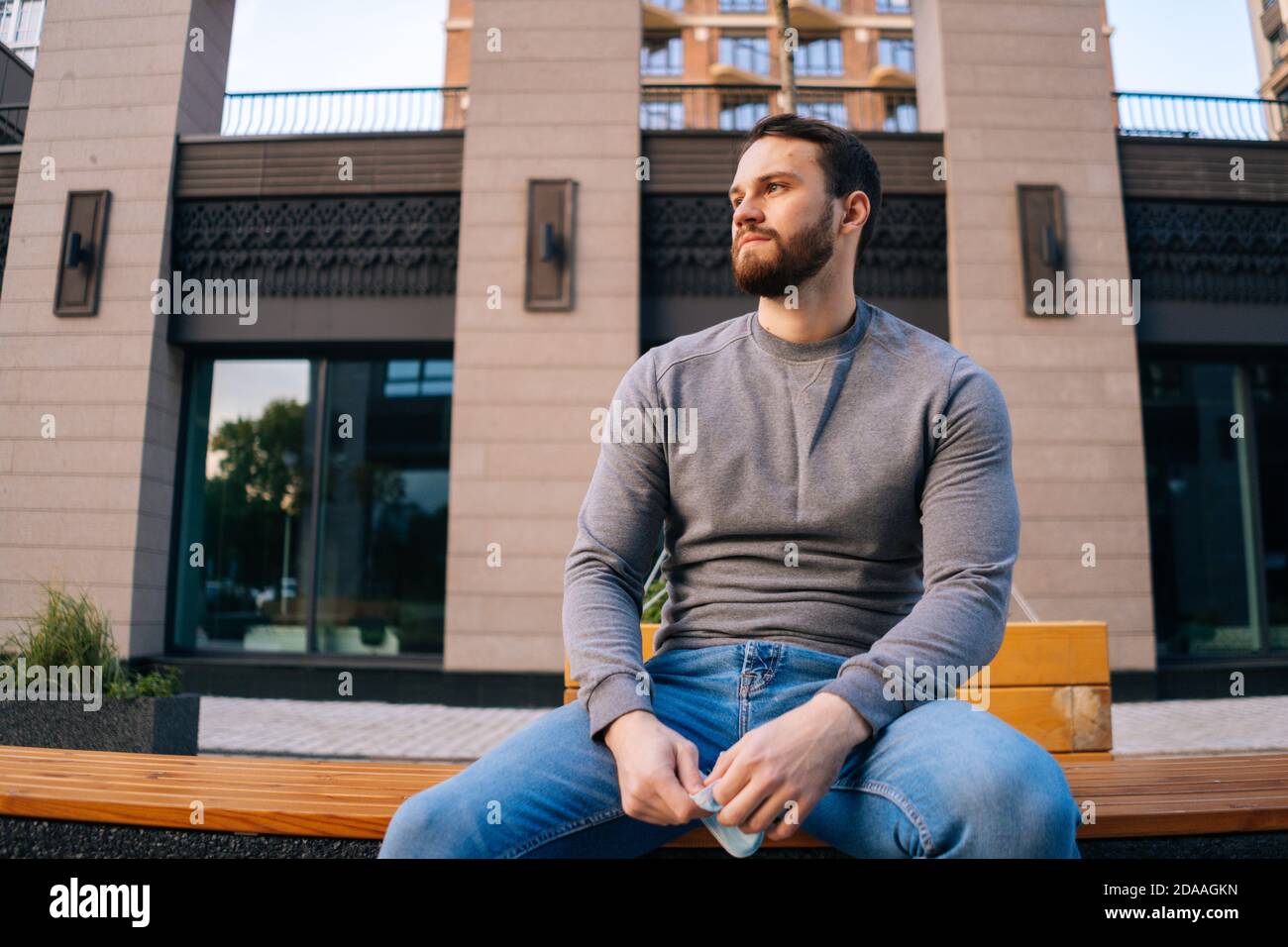 Low-angle shot of upset young man sitting on urban bench at empty European city street. Stock Photo