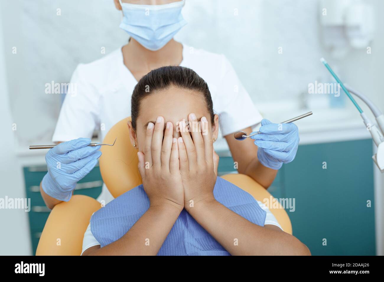 Scared little latin girl in dental chair covers face and does not give young woman doctor examination teeth in clinic Stock Photo