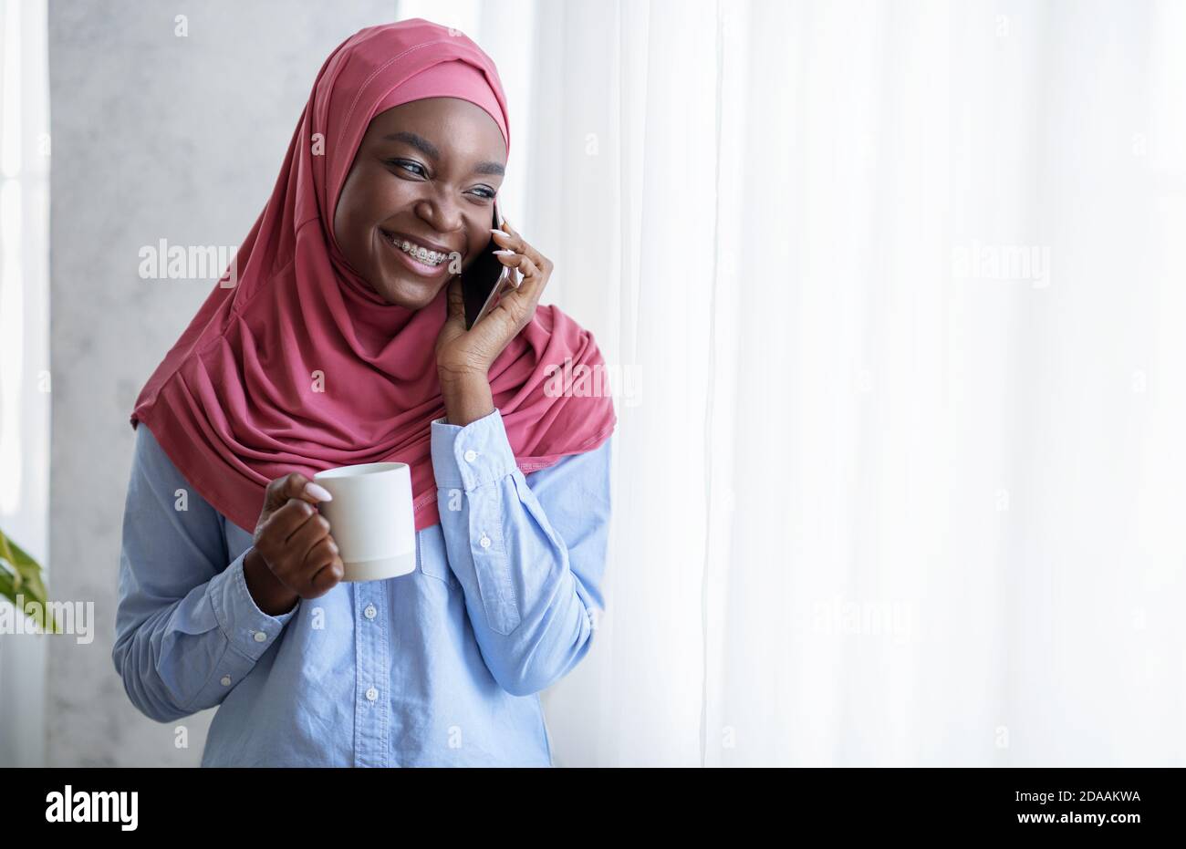 Smiling Black Muslim Lady Relaxing Near Window With Cellphone And ...