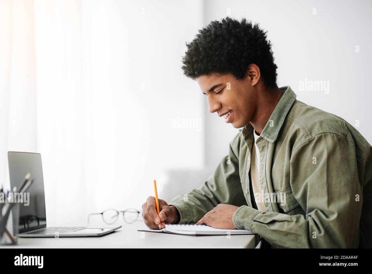 Web-based training concept. Black male student writing in notebook in front of laptop at home, empty space Stock Photo