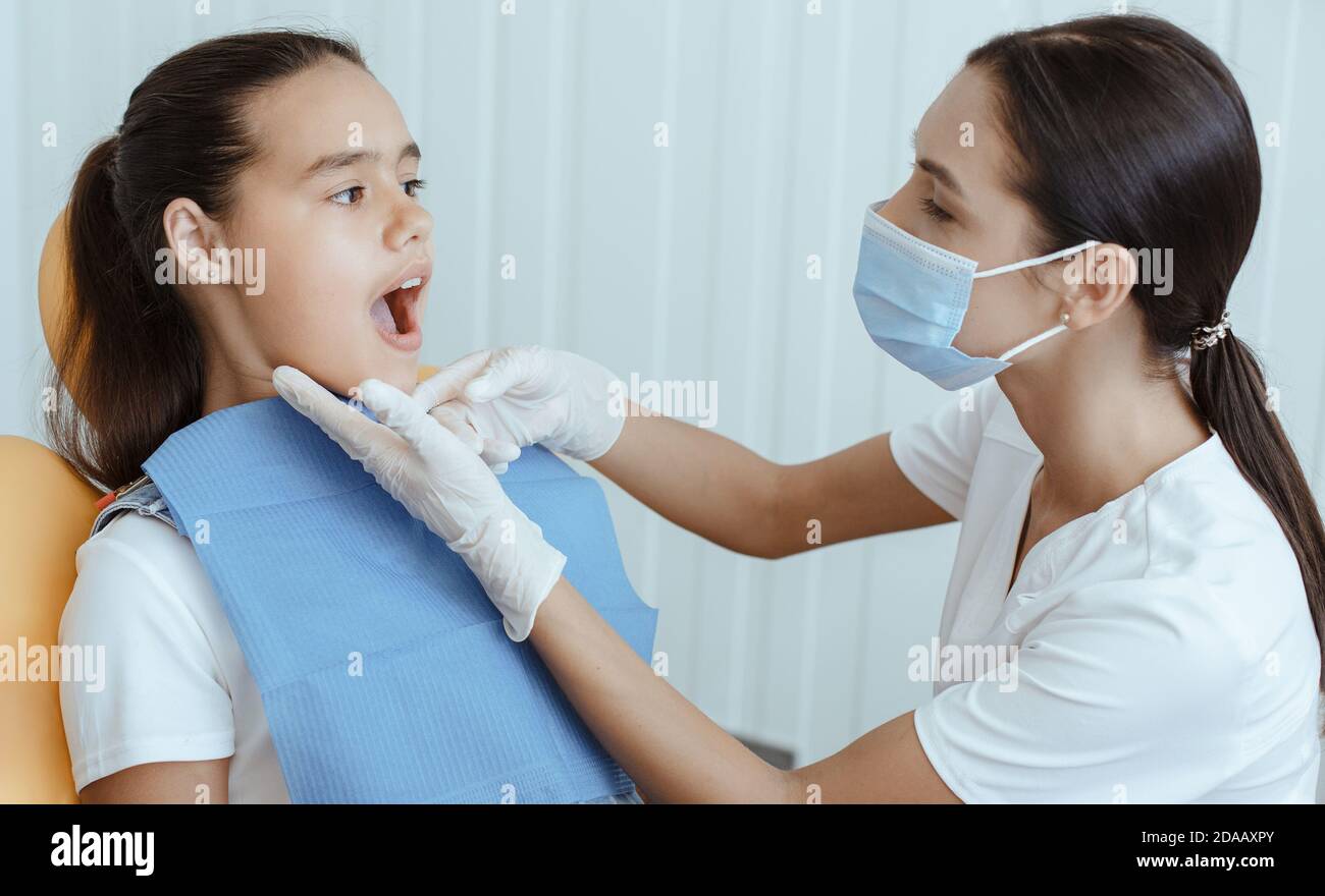 Millennial lady doctor in uniform, protective mask and rubber gloves examines of small child with open mouth Stock Photo