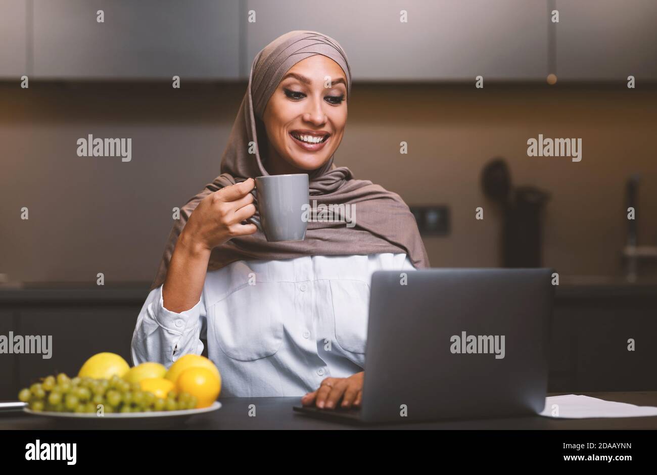 Muslim Woman At Laptop Watching Movie Drinking Coffee In Kitchen Stock ...