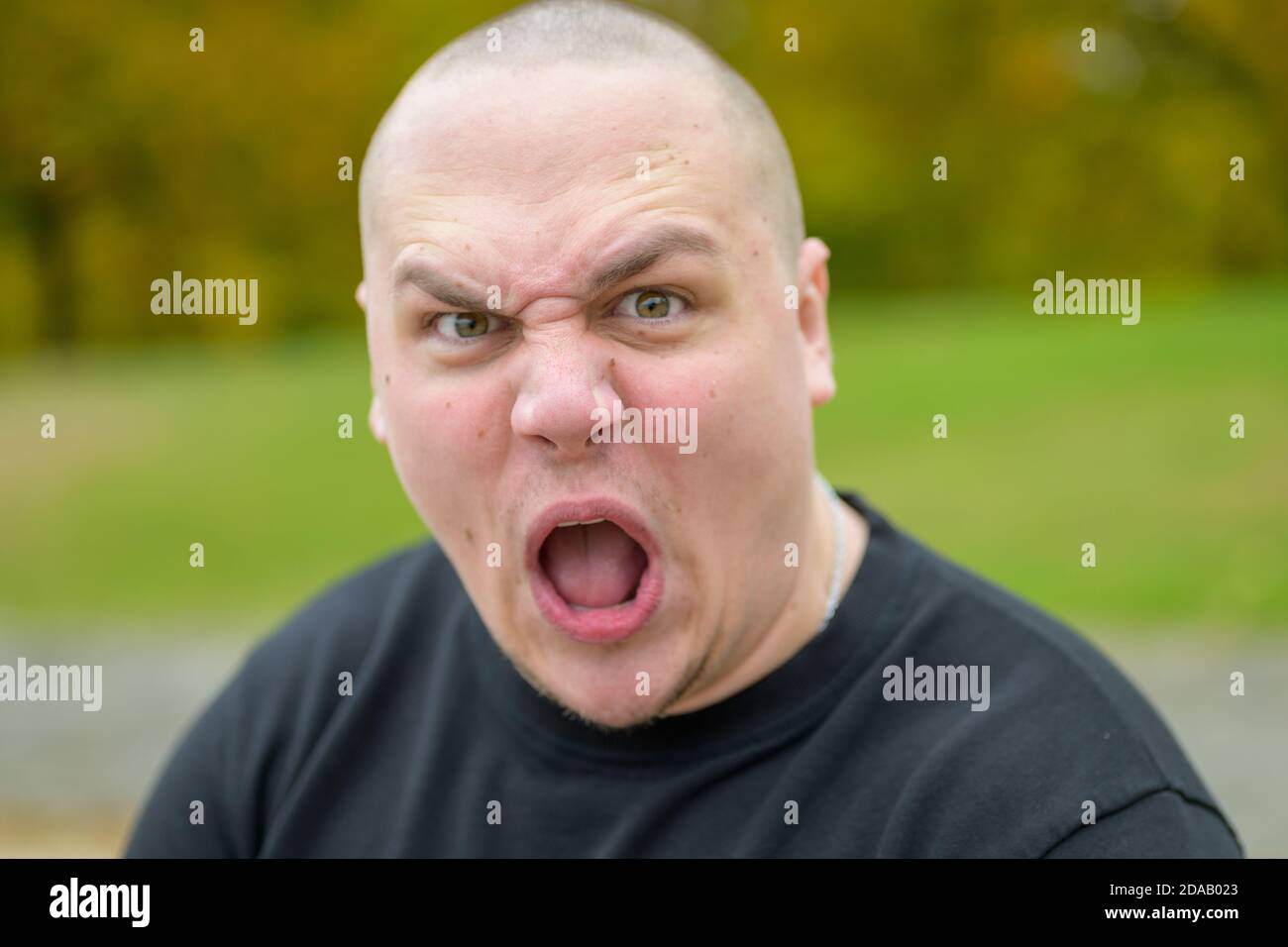 Young man pulling a menacing ugly face at the camera gawping and frowning with a baleful stare outdoors in a park Stock Photo